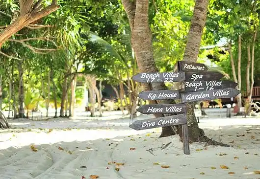 Rustic wooden signpost on a sandy path at Pom Pom Island, directing guests to the restaurant, spa, dive centre, and various luxury villas.