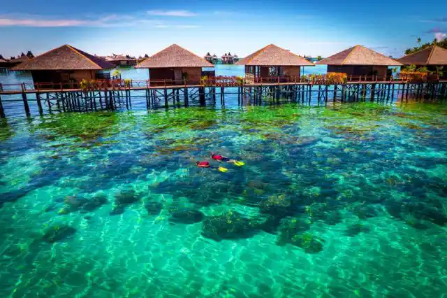 Snorkelers exploring the vibrant coral reef in crystal-clear turquoise water directly in front of the overwater chalets at Sipadan Water Village.