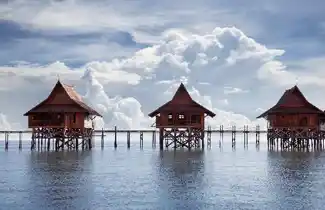 Iconic overwater chalets of the Kapalai Dive Resort lined up perfectly over the calm, crystal-clear turquoise sea under a blue sky with fluffy clouds.