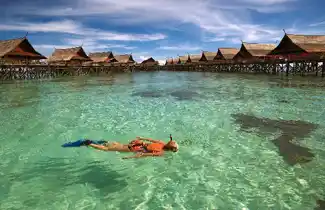 Snorkeler floating effortlessly in the crystal-clear turquoise waters of Kapalai, with the resort's beautiful overwater chalets in the background.
