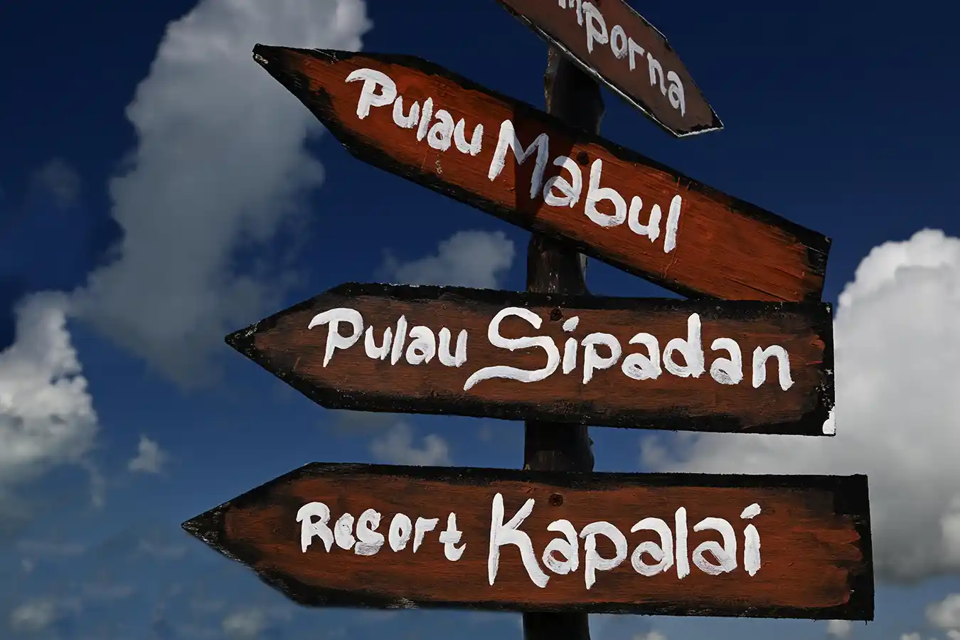 Rustic wooden signpost pointing the way to the legendary diving destinations of Pulau Sipadan, Pulau Mabul, and Resort Kapalai against a beautiful blue sky.
