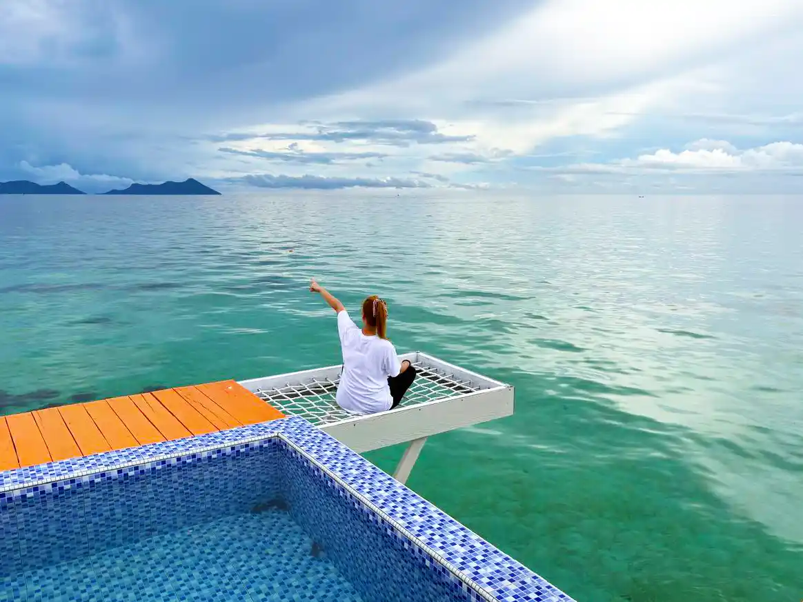 Guest relaxing on an over-water net hammock at Danglai Resort, pointing towards the beautiful view of Bohey Dulang island.