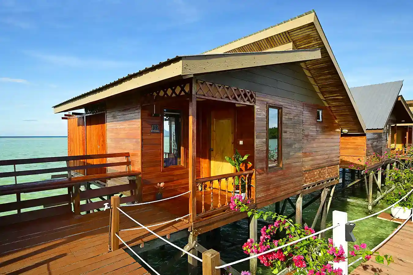 Wooden boardwalk connecting the beautiful overwater chalets at Larapan Water Village, decorated with vibrant pink bougainvillea.