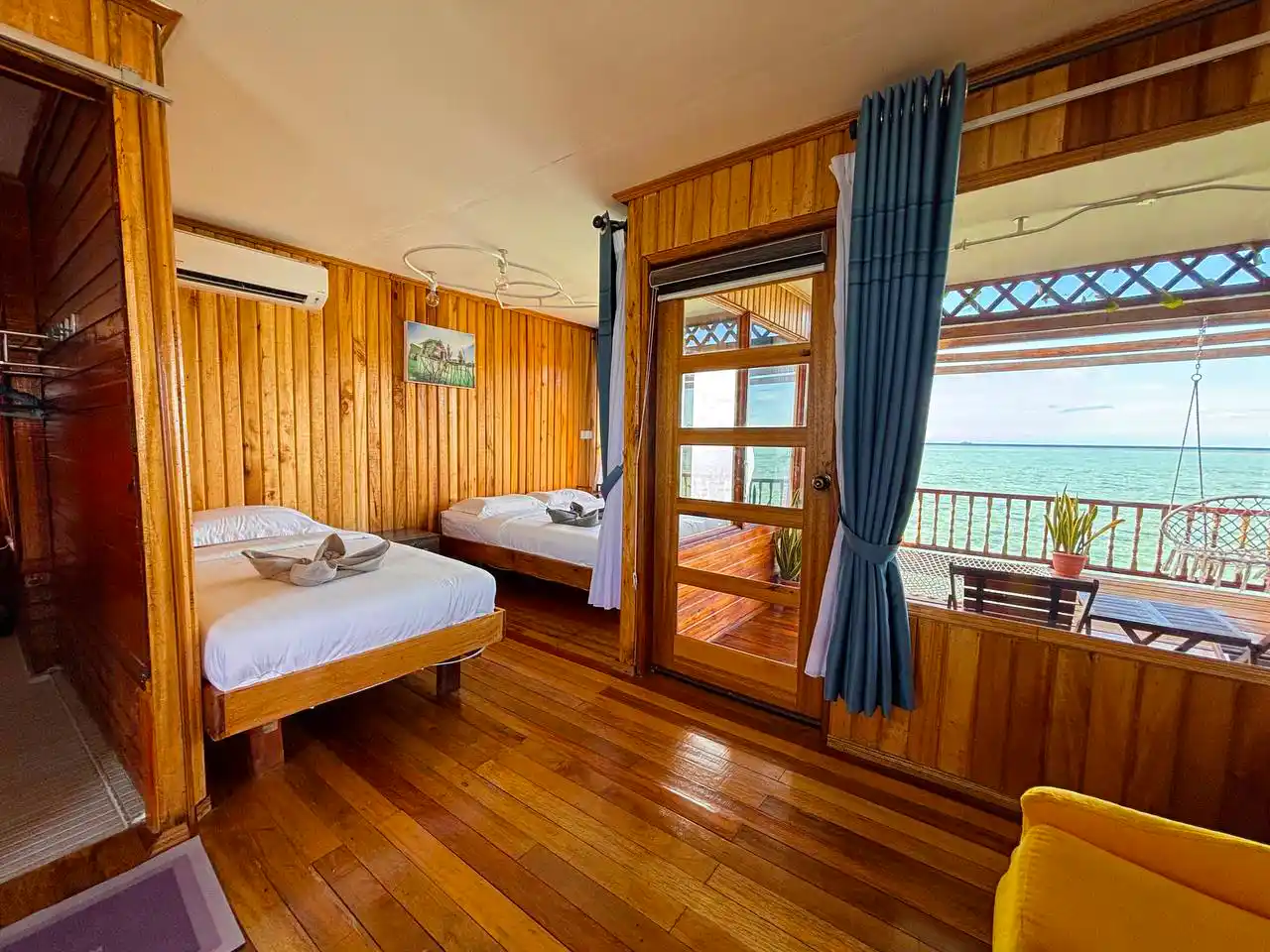 Spacious and clean wood-paneled room at Larapan Water Village, with two beds and a glass door opening onto a private balcony with a sea view.