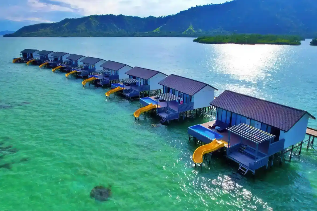 Spectacular drone view of the row of modern overwater chalets at Sky Resort Semporna, with the lush mountains of Timbun Mata island in the background.
